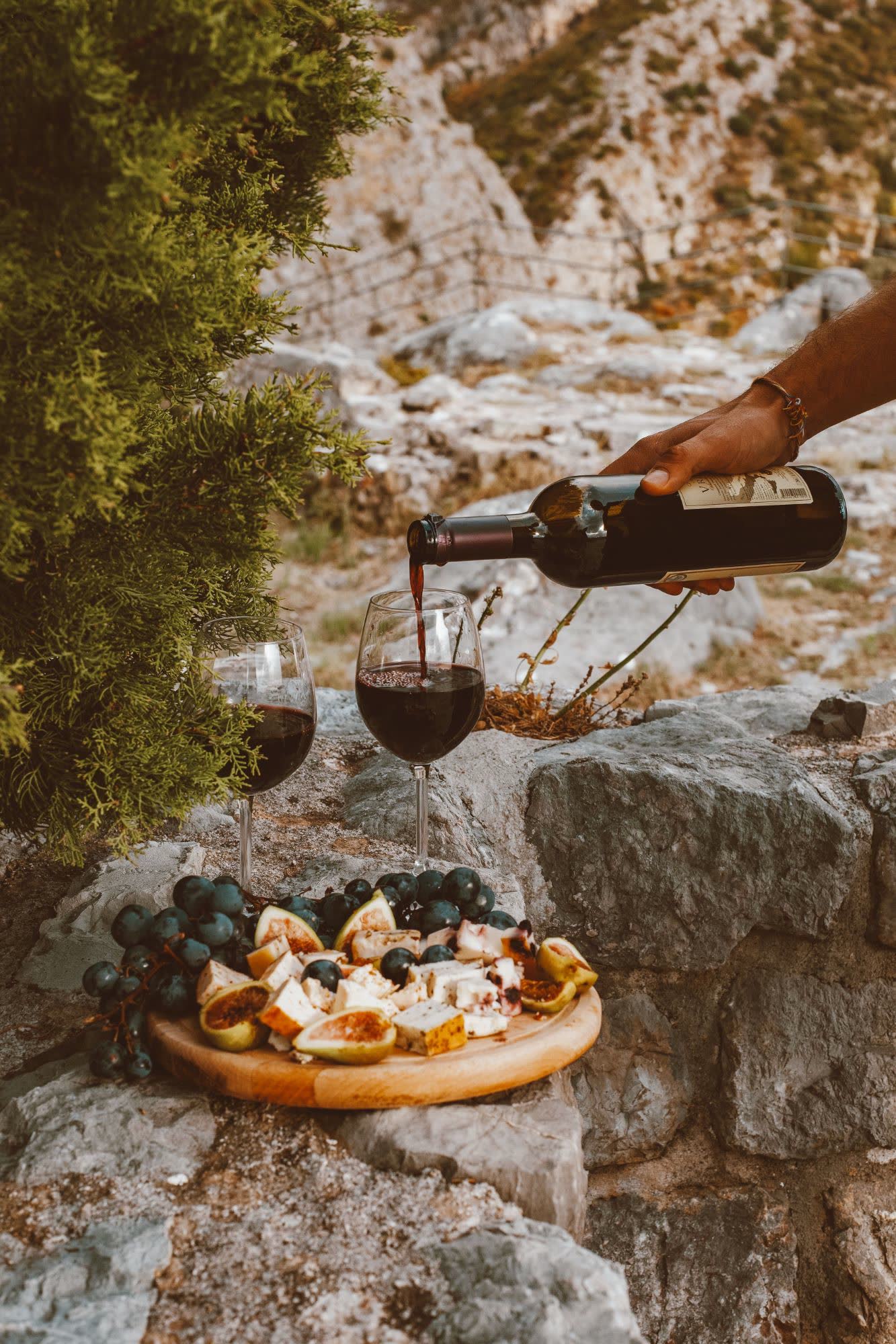 A hand pouring red wine beside a board of figs, grapes and cheese on a stone wall above a rocky hillside